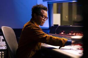 afrikanisch amerikanisch Musik- Produzent betreibt Audio- Konsole mit ziehen um Fader und Meter, Aufzeichnung Spuren im Steuerung Zimmer beim Studio. jung Klang Designer produzieren Lieder zum ein Neu Album. foto