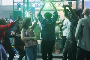 aktiv Menschen Tanzen und erziehen Hände während Party machen im Nachtclub. jung Männer und Frauen ziehen um zu elektronisch Musik- schlägt und entspannend auf überfüllt Tanzfläche beim Diskothek foto