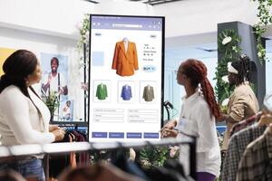 Geschäft Assistent Portion Klient mit Kiosk Service, weiblich Mitarbeiter geben Hilfe im Kleidung speichern. Käufer suchen zum modisch Artikel auf interaktiv Tafel Monitor, selbst Bestellung. foto