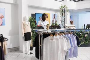 Einkaufen Einkaufszentrum afrikanisch amerikanisch Assistent Arbeiten im Mode speichern, Verlegung aus Kleider zum Verkauf. Ausstellungsraum Mann Mitarbeiter Überprüfung Fan-Shop und Anzeigen Kleidungsstück auf Kleiderbügel zum Kunden foto