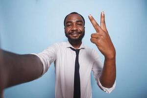 pov von jung männlich Unternehmer Herstellung online vlog zum Sozial Medien Werbung. schwarz Geschäftsmann nehmen ein Selfie mit Frieden Zeichen Hand Geste im Vorderseite von isoliert Blau Hintergrund. foto