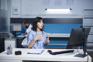 weiblich Krankenschwester im ein Krankenhaus kommuniziert durch ein Anruf mit ihr Desktop Stk. kaukasisch Frau sitzend im das Klinik Büro haben ein virtuell treffen. Zusammenarbeit und Technologie im Gesundheitspflege. foto