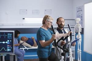 medizinisch Spezialist diskutieren Physiotherapie mit Rentner Wer ist Reiten Fahrrad zu heilen von muskulös Verletzung. Alten Dame im physisch Behandlung Center mit stationär Fahrrad zum Erholung. foto