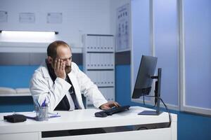 Bild präsentieren ein müde kaukasisch Arzt sitzend beim ein Schreibtisch mit ein Desktop Computer nach ein lange Tag im Krankenhaus. erschöpft männlich Arzt berühren seine Gesicht im ein Klinik Büro Zimmer. foto