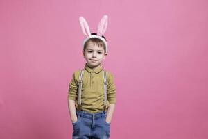 jung froh Kind tragen süß Hase Ohren im Vorderseite von Kamera, Gefühl aufgeregt Über Ostern Veranstaltung Fest und Stehen gegen Rosa Hintergrund. heiter klein Junge mit bezaubernd Outfit. foto