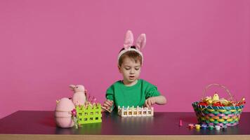 glücklich Kleinkind mit Hase Ohren arrangieren Korb gefüllt mit gemalt Eier, Erstellen festlich Vereinbarungen zum das Ostern Sonntag Feier. heiter wenig Kind genießt Kunst und Handwerk. Kamera b. foto