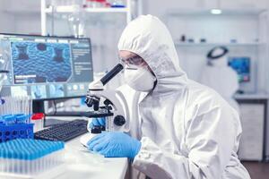 Arzt recherchieren und auswerten Virus mit Mikroskop im Labor tragen PSA. Wissenschaftler im schützend passen Sitzung beim Arbeitsplatz mit modern medizinisch Technologie während global Epidemie. foto