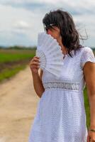 Frau ist halten ein Weiß Ventilator im ihr Hand während Gehen auf ein Schmutz Straße foto