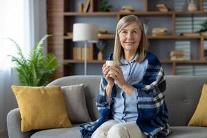 ein entspannend Szene von ein Frau Sitzung auf ein Couch, bedeckt mit ein Decke, und halten ein Kaffee Becher im ein gut dekoriert Leben Zimmer. foto