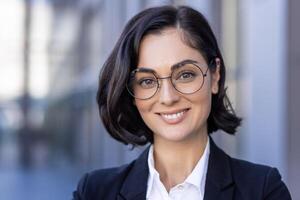 Nahansicht Foto von ein jung schön erfolgreich weiblich Anwalt im ein passen und Brille lächelnd beim das Kamera und Stehen außen.