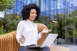 ein jung afrikanisch amerikanisch Geschäftsfrau sitzt auf ein Bank in der Nähe von ein Büro Center und Gespräche auf ein Anruf auf ein Laptop auf ihr Schoß. foto