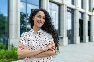 lächelnd jung Fachmann spanisch Frau mit lockig Haar Stehen selbstbewusst draußen ein Büro Gebäude auf ein sonnig Tag. foto