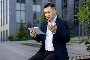 verärgert und besorgt jung asiatisch männlich Geschäftsmann Sitzung auf ein Bank draußen ein Büro Gebäude tragen Kopfhörer ,Und suchen enttäuscht beim ein Tablette im seine Hand. foto