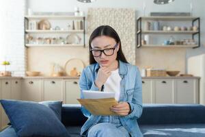 ein besorgt jung asiatisch Frau empfangen ein Brief mit Rechnungen zum Kredit, mieten, Hypothek. Sitzung beim Zuhause auf das Couch, halten ein Briefumschlag mit ein Nachricht, Quittungen und Schulden. foto