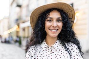 schön jung Latein amerikanisch Frau Porträt, Frau Gehen im Abend Stadt im Hut mit lockig Haar im warm Wetter, lächelnd und suchen beim Kamera schließen hoch. foto