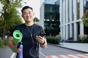 Nahansicht Porträt von ein jung asiatisch männlich Sport Trainer und Lehrer Stehen draußen im Kopfhörer, halten ein Telefon und ein Matte, lächelnd beim das Kamera. foto