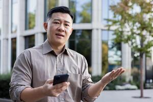 Nahansicht Porträt von ein traurig jung asiatisch Mann Stehen in der Nähe von ein Büro Gebäude mit ein Telefon im seine Hände und suchen besorgt beim das Kamera, werfen oben seine Hände im Frustration. foto