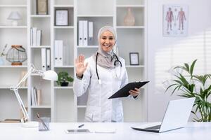lächelnd Muslim Arzt im Hijab Gruß mit ein Welle während halten ein Zwischenablage im ein gut beleuchtet medizinisch Büro. foto