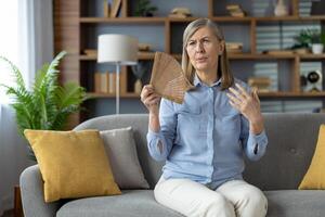 genervt Senior weiblich mit Hand Ventilator und Leiden von Sommer- Überhitzung und Abwesenheit von Klimaanlage. erschöpft Frau Sitzung auf Couch beim Leben Zimmer während Gefühl Beschwerden und Mangel von frisch Luft. foto