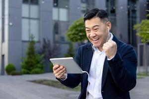 Nahansicht Foto von ein jung asiatisch Mann im ein Geschäft passen Sitzung draußen das Büro tragen Kopfhörer, suchen beim ein Bildschirm Tablette und Glücklich, zeigen ein Sieg Geste mit seine Hand.