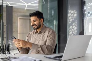 ein heiter jung Geschäftsmann engagiert mit seine Smartphone, sitzend im ein gut beleuchtet, zeitgenössisch Büro Raum, präsentieren ein Mischung von Technologie und beiläufig korporativ Stil. foto