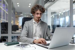 Fachmann jung Geschäftsmann mit lockig Haar fleißig Arbeiten auf ein Laptop im ein zeitgenössisch Büro Raum. foto