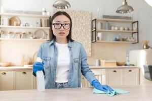 Porträt von ein jung asiatisch Hausfrau Reinigung beim heim. Tücher das Tabelle im das Küche mit ein Lappen im Gummi Handschuhe, Verwendet Haushalt Chemikalien. er ist Stehen, halten ein sprühen, suchen beim das Kamera. foto