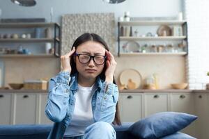 ein jung krank asiatisch Frau sitzt auf das Sofa beim heim, das Frau hat ein Kopfschmerzen, ein Nahansicht Foto von ein Schüler im Brille