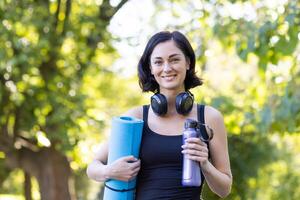 ein lächelnd Frau ausgestattet mit Yoga Matte, Kopfhörer, und Wasser Flasche genießen das natürlich draussen Rahmen zum ihr Fitness Routine. foto