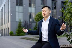 ernst asiatisch männlich Geschäftsmann Sitzung auf Bank in der Nähe von Arbeiten Büro im Lotus Position und meditieren mit geschlossen Augen. foto