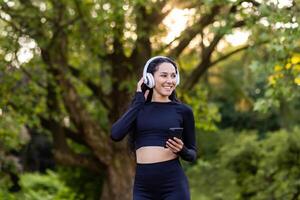 erfolgreich schlank Latein amerikanisch Frau im Park während aktiv Übung und Joggen Verwendet Smartphone Anwendung, Sportlerin lächelnd im Kopfhörer Hören zu Audio- Podcasts und online Radio. foto