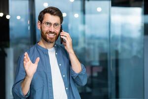 Porträt von ein reifen erfolgreich Geschäftsmann Innerhalb das Büro, das Mann ist lächelnd und suchen beim das Kamera durch das Fenster während Stehen und reden fröhlich und lächelnd auf das Telefon. foto