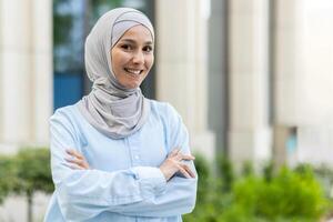 Porträt von ein heiter Muslim Frau tragen ein Kopftuch, selbstbewusst Stehen mit ihr Waffen gekreuzt im ein städtisch Einstellung. foto