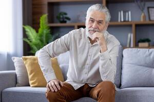 ein Alten Mann mit ein stilvoll Bart und Weiß Haar sitzt nachdenklich auf ein Couch, seine Hand auf seine kinn, porträtieren ein heiter Moment im ein modern Leben Zimmer. foto