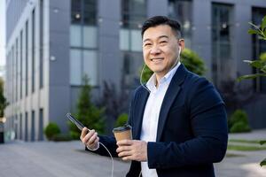 Porträt von ein asiatisch jung männlich Büro Arbeiter tragen Kopfhörer und halten ein Telefon und ein Tasse von Kaffee ruhen Sitzung draußen ein Büro ,Center und lächelnd beim das Kamera. foto