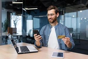 heiter und lächelnd bärtig Geschäftsmann tun online Einkaufen und Bank Geld überweisen, Freiberufler halten Bank Anerkennung Karte und Smartphone, Mann Sitzung beim Schreibtisch Innerhalb Büro. foto