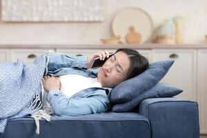 ein krank asiatisch Frau beim Zuhause ist Lügen auf das Sofa unter ein Decke, sie hat ein stark Bauch schmerzen, das Frau Anrufe das Arzt und fragt zum Hilfe foto