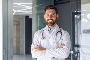 Nahansicht Porträt von ein lächelnd jung männlich Arzt, Assistent, Schüler Stehen im das Krankenhaus Büro und selbstbewusst suchen beim das Kamera mit seine Waffen gekreuzt auf seine Brust. foto