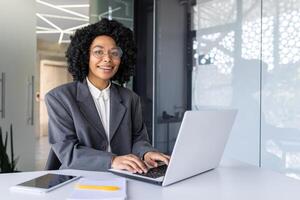 Porträt von jung schön Geschäft Frau im Geschäft Anzug, afrikanisch amerikanisch Frau lächelnd und suchen beim Kamera, weiblich Arbeiter mit Laptop Sitzung beim Tabelle Innerhalb Büro. foto