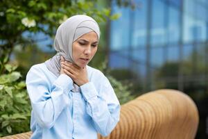 besorgt Frau im Hijab Gefühl Reizung im Kehle und erleben Symptome von Grippe Virus während Gehen Nieder Straße. krank weiblich berühren wund Stelle auf Körper und Denken Über verschreiben Behandlung. foto