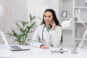 ein heiter weiblich Arzt mit lockig Haar Gespräche auf das Telefon während Sitzung beim ihr Schreibtisch im ein gut beleuchtet Büro, umgeben durch medizinisch und Büro Lieferungen. foto