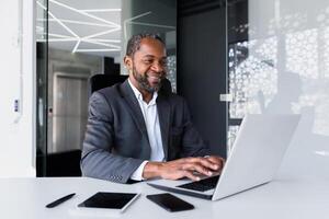 erfolgreich afrikanisch amerikanisch Manager Innerhalb Büro beim Arbeitsplatz, Mann Arbeiten mit Laptop lächelnd und glücklich mit Leistung Ergebnisse und finanziell Geld, Boss im Geschäft passen Tippen auf Klaviatur. foto