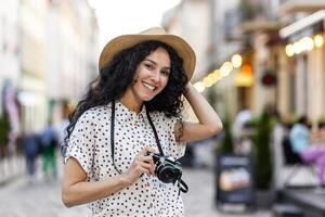 Porträt von jung schön Frau Tourist mit Kamera, spanisch Frau mit lockig Haar im Hut Gehen im Abend Stadt lächelnd und suchen beim Kamera schließen hoch. foto