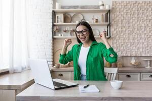 ein jung glücklich Frau sitzt beim Zuhause mit ein Laptop, empfangen gut Nachricht, bestanden das Prüfung, finanziell dazugewinnen foto