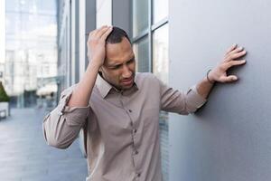 krank afrikanisch amerikanisch Mann draußen modern Büro Gebäude, Mann hat stark Kopfschmerzen und Migräne, Arbeiter im Hemd auf brechen übermüdet foto