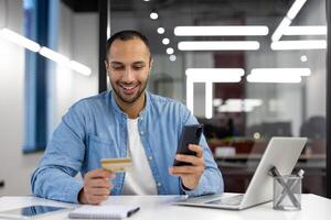 ein Mann ist lächelnd während mit seine Zelle Telefon zu Zahlen zum etwas mit ein Anerkennung Karte. Konzept von Bequemlichkeit und Leichtigkeit im Herstellung Transaktionen mit Technologie foto