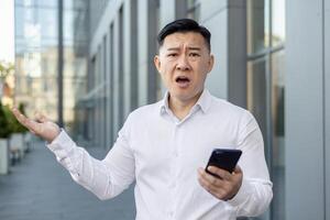 Nahansicht Porträt von ein wütend jung asiatisch Mann im ein Weiß Hemd Stehen draußen ein Büro Gebäude, halten ein Telefon und werfen seine Hände oben im Frustration und suchen beim das Kamera. foto