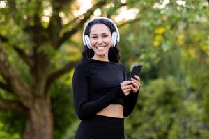 aktiv Brünette Frau tragen kabellos Headset und schwarz Sportbekleidung während suchen beim Kamera mit Smartphone im Hand. lächelnd weiblich tun Täglich Übungen zu Musik- auf Hintergrund von Öffentlichkeit Park. foto