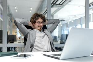 ein jung zuversichtlich jung Mann sitzt im das Büro beim das Schreibtisch, sieht aus zufrieden beim das Laptop Bildschirm, ruht mit seine Hände hinter seine Kopf. foto