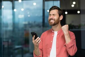 ein erfolgreich Geschäftsmann freudig feiert Sieg und gut Erfolge, ein Mann im ein rot Hemd Innerhalb das Büro empfangen ein online Benachrichtigung, halten ein Smartphone im seine Hände foto